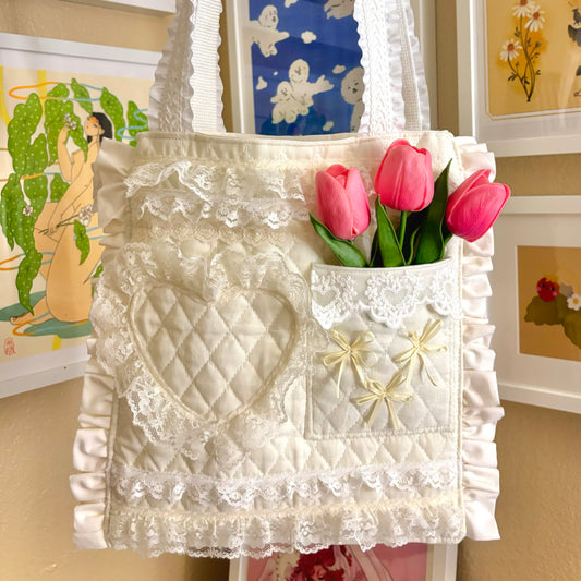 A white ruffle lace tote bag with a quilted pocket, displayed against a framed artwork background. Pink tulips are in the quilted pocket.