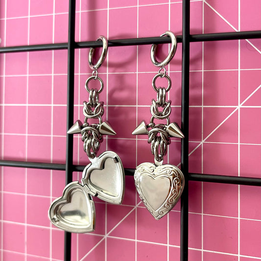 A pair of heart-shaped locket chainmaille earrings with spikes on a pink grid background
