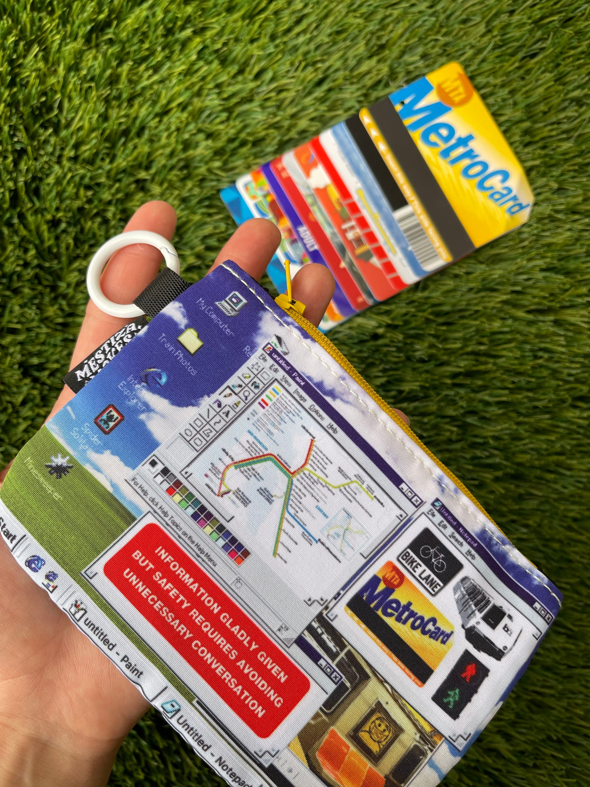 A zippered pouch featuring a detailed Windows desktop-inspired graphic print, held up with artificial grass as the background. The pouch features a yellow zipper, a white carabiner ring, and a black brand tag reading 'Mestiza Makes'. A row of Metro Cards lies on green artificial turf in the background.