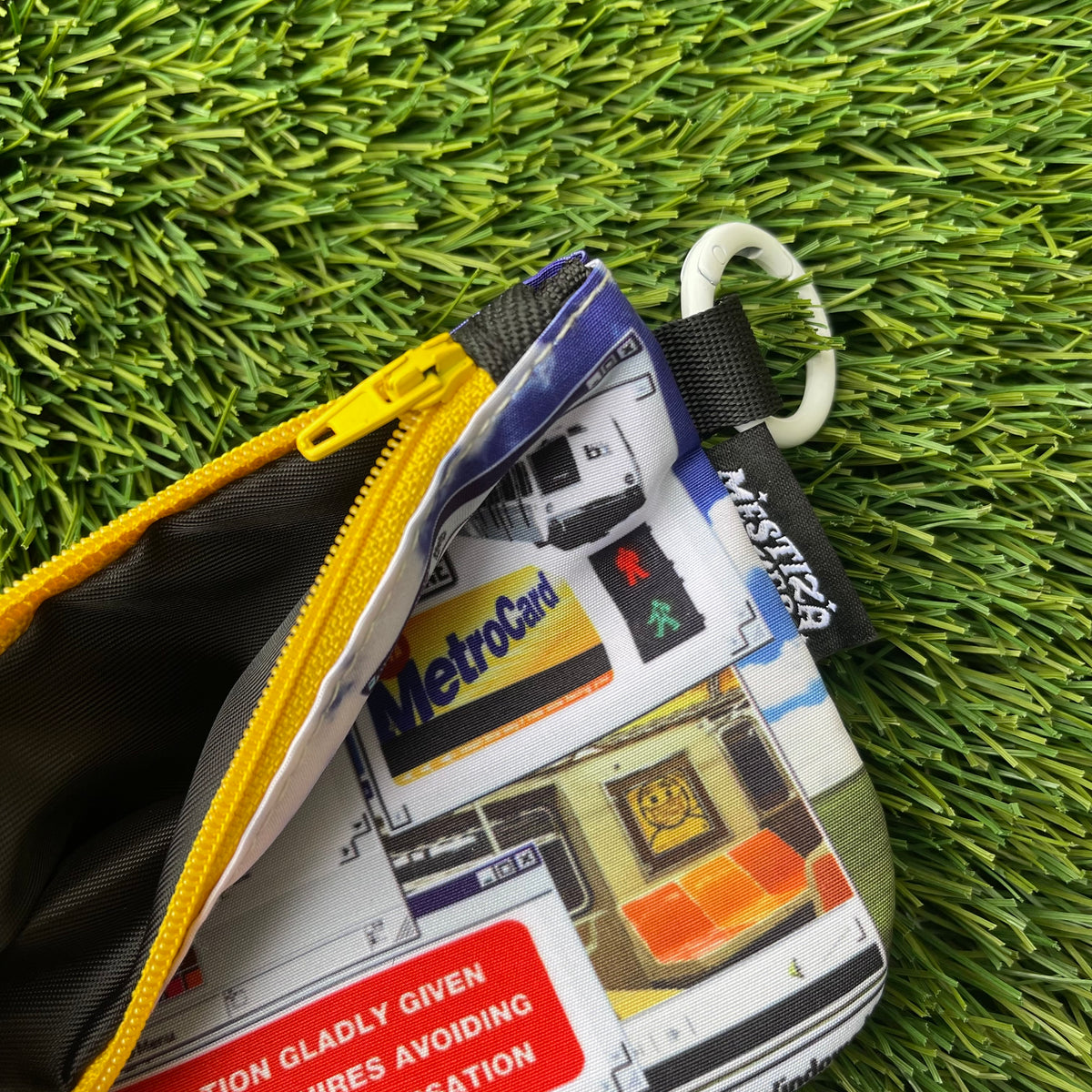 A close up photo of a zippered pouch with a detailed Windows desktop-inspired graphic print, laid flat on artificial green grass. The pouch features a yellow zipper, a white carabiner ring, and a black brand tag reading 'Mestiza Makes'.