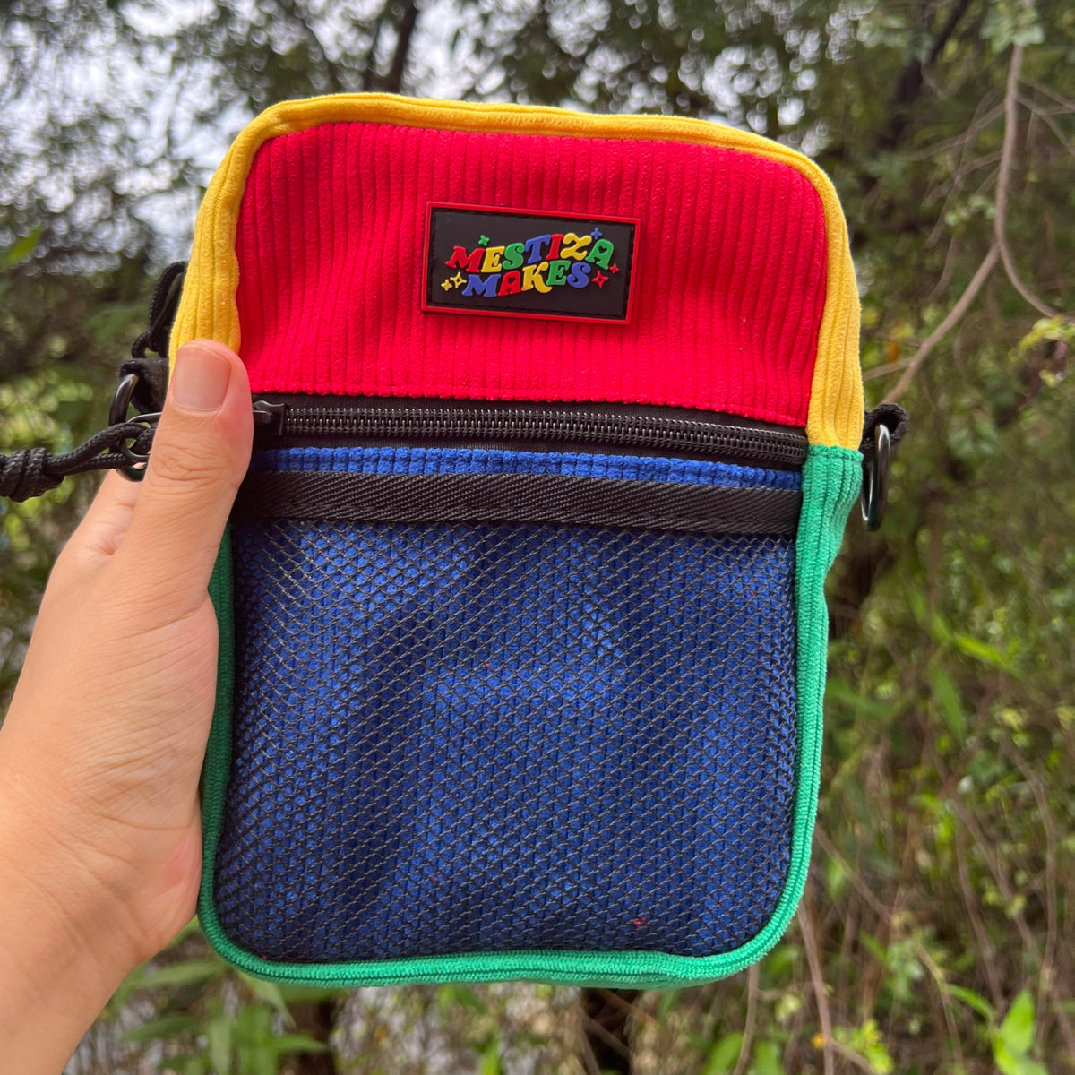 A colorblock sling bag with red, yellow, blue, and green corduroy exterior held in a person's hand, featuring a front mesh pocket and an adjustable strap.