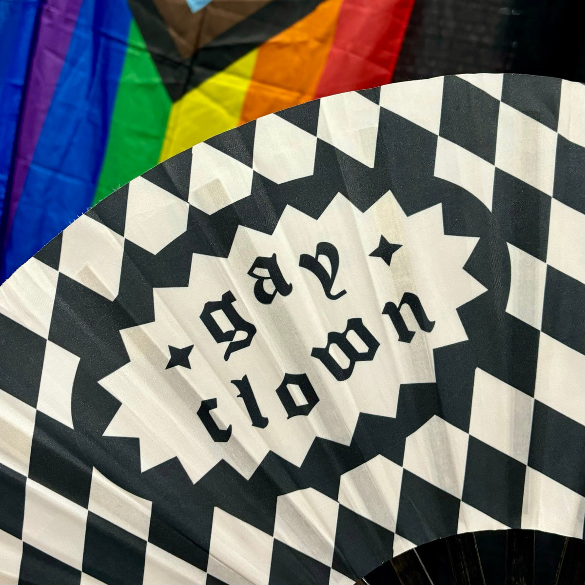 A bamboo fan with a black and white checkered harlequin pattern, featuring the phrase 'GAY CLOWN' in lowercase gothic lettering.