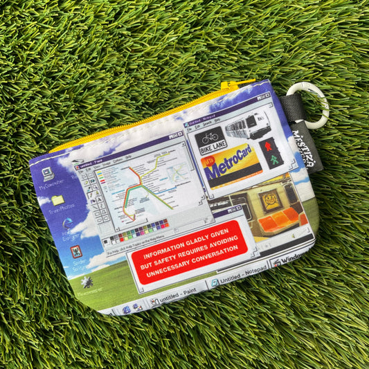 A zippered pouch with a detailed Windows desktop-inspired graphic print, laid flat on artificial green grass. The pouch features a yellow zipper, a white carabiner ring, and a black brand tag reading 'Mestiza Makes'.