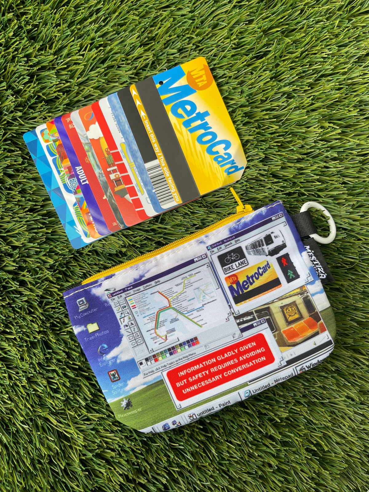 A zippered pouch with a detailed Windows desktop-inspired graphic print, laid flat on artificial green grass. The pouch features a yellow zipper, a white carabiner ring, and a black brand tag reading 'Mestiza Makes'. There are a row of public transit cards placed above the pouch. 