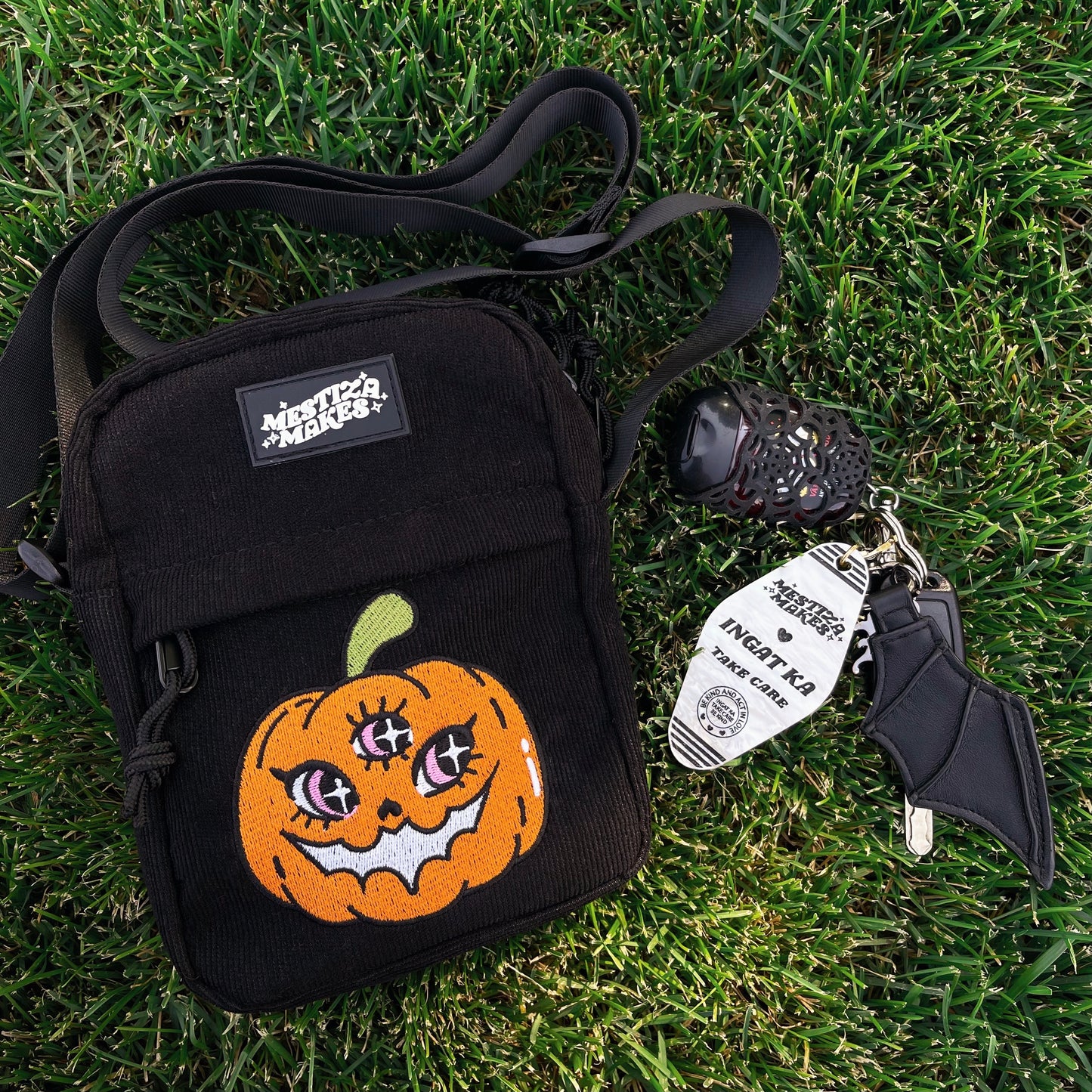 Black corduroy crossbody bag with an embroidered pumpkin design on the front, displayed on a grassy background with a bat and bat mouth accessory.